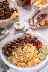 A hand holding a fork over grilled beef kebab with colorful basmati rice, roasted tomato, and cucumber on a white plate, surrounded by yogurt dip, salad, and bread on a marble surface