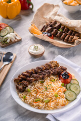 Top view of grilled beef kebab with colorful basmati rice, roasted tomato, cucumber slices, yogurt dip, fresh vegetables, and pita bread on marble surface with wooden cutlery tray beside