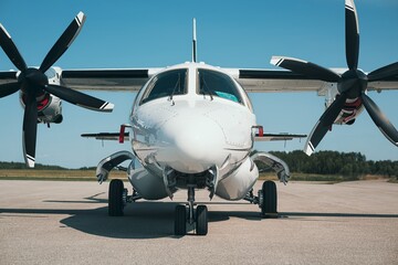 Front view of a modern twin turboprop aircraft parked on a sunlit airport tarmac. Visible are the spinning propellers, landing gear, cockpit windows, and sleek fuselage. 