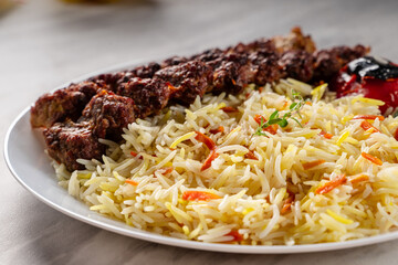 Close-up of grilled beef kebab served with colorful basmati rice and roasted tomato on a white ceramic plate, placed on a light marble surface under soft natural lighting