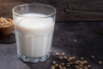 A glass of fresh soy milk placed on a dark stone surface with scattered soybeans and a wooden bowl in the background, highlighting plant-based nutrition in natural rustic lighting