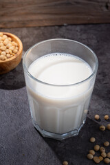 A glass of fresh soy milk placed on a dark stone surface with scattered soybeans and a wooden bowl...