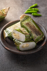 Traditional Indonesian lemper made of sticky rice filled with shredded chicken, wrapped in grilled banana leaves, served on a brown clay plate with green chilies over a dark stone surface