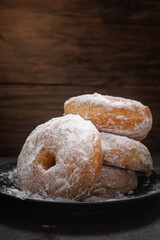Close-up of stack of sugar-coated donuts on a black plate, with powdered sugar scattered around, placed on dark stone surface and wooden background, highlighting texture and sweetness