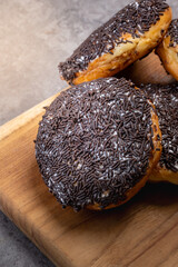 Close-up of chocolate sprinkle donuts stacked on a wooden cutting board over a gray stone background, with white icing and textured topping, presenting a rustic and indulgent dessert look