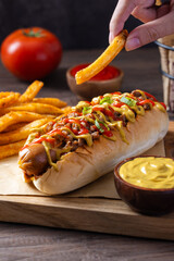 A hand holding a seasoned French fry near a loaded hot dog topped with meat, mustard, ketchup, and scallions, served with more fries and dipping sauces on rustic wooden table in natural light