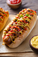 Close-up of a loaded hot dog with sausage, ground meat, mustard, ketchup, and green onion topping on a wooden board, with tomato and sauces in the background over rustic wooden surface