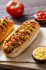 Close-up of a loaded hot dog with sausage, ground meat, mustard, ketchup, and green onion topping on a wooden board, with tomato and sauces in the background over rustic wooden surface