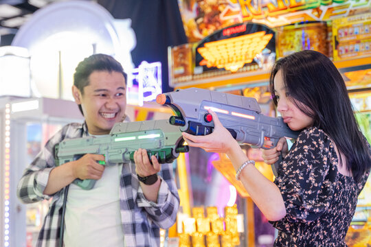 Young Asian couple playing a laser shooting arcade game, aiming toy guns at each other with playful and competitive expressions, surrounded by colorful neon-lit machines in a lively game center
