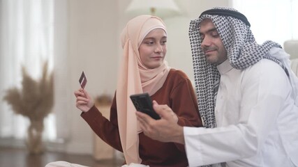 Muslim couple using credit card for online shopping holding smartphone at home, concept of cashback discount spending islamic lifestyle payment, e-wallet mobile banking, halal e-commerce platform - Powered by Adobe