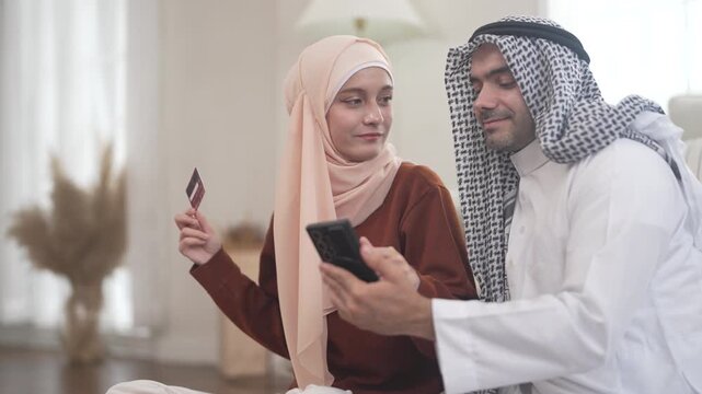 Muslim couple using credit card for online shopping holding smartphone at home, concept of cashback discount spending islamic lifestyle payment, e-wallet mobile banking, halal e-commerce platform