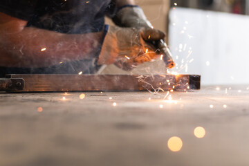An adult male works in an old workshop. He holds a welding machine in his hand and welds the metal structure