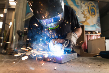An adult male works in an old workshop. He holds a welding machine in his hand and welds the metal structure