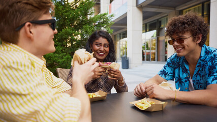 Three young friends eating burgers and fries at outdoor table. Concept of street food advertising, casual social moments, youth lifestyle promotion, delivery platform visuals.
