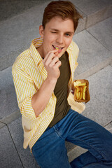 Young man winking while eating fries on stone steps outside in casual clothing. Concept of playful...
