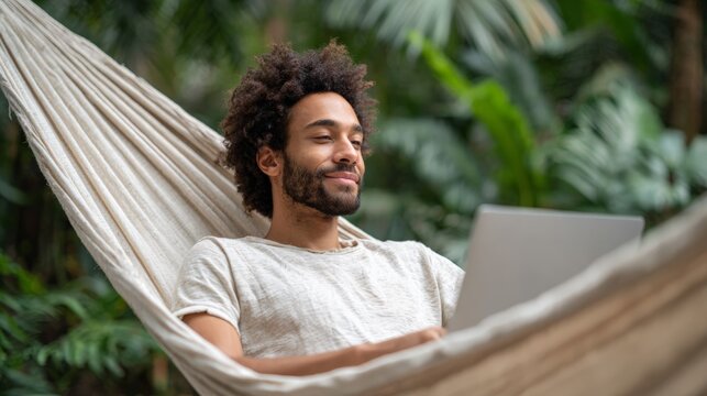 Young freelancer working remotely using laptop while relaxing in hammock surrounded by lush tropical plants, enjoying work and travel lifestyle