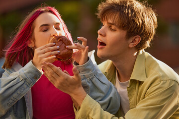 Obraz premium Young woman taking huge bite of burger while young man reacts with surprise. Concept of competitive eating, funny youth behavior, social sharing, food delivery visuals.