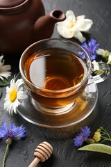 Aromatic herbal tea in glass cup, different flowers and teapot on black table, closeup