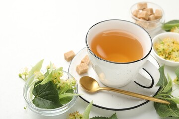 Tasty linden tea in cup, spoon, sugar cubes, leaves and flowers on white table, closeup