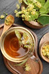 Tasty linden tea in cup, spoon, leaves and flowers on dark textured table, flat lay