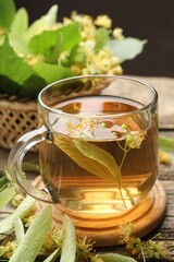 Tasty linden tea in cup, leaves and flowers on wooden table against dark background, closeup