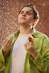 Young man savoring last bite of burger under refreshing water spray. Concept of food obsession,...