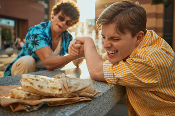 Two young men arm wrestling for wraps outdoors on summer day. Concept of friendly competition, food...