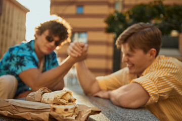 Two friends engaged in arm wrestling outside centered around street food. Concept of food as prize, lifestyle visual for youth branding, fast food motivation and delivery culture.