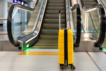 A yellow suitcase stands near an escalator inside an airport terminal. Journey, travel, movement...
