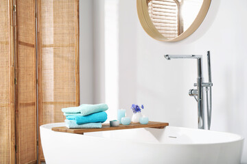 Wooden board with soft towels, sea salt, flowers and burning candles on tub in bathroom