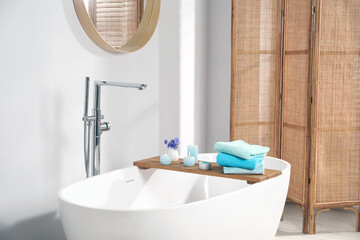 Wooden board with soft towels, sea salt, flowers and burning candles on tub in bathroom