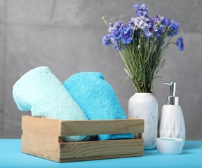Soft towels, liquid soap, sea salt and flowers in wooden basket on light blue table