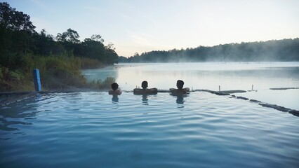 Tourists relaxing in the hot spring of Talaga Bodas, Garut, Indonesia, enjoying the serene volcanic lake at sunrise with mist drifting above the water.