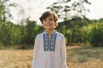 A young boy smiles confidently while standing in a sunlit field, wearing a traditional embroidered...