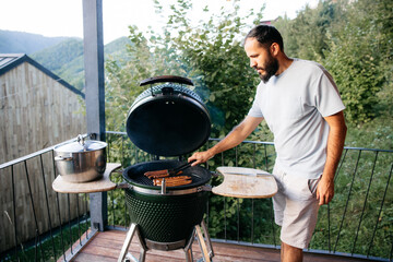Man grilling sausages on modern green ceramic grill on balcony with mountain view