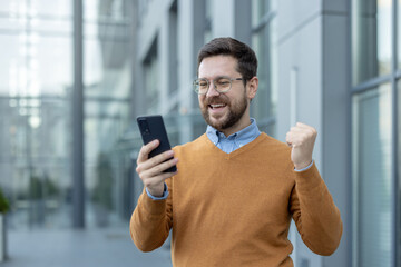 Close-up shot of a happy young man standing on the street, looking at the phone screen and...