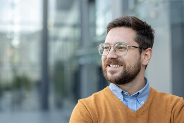 Close-up photo of a young businessman with a beard and glasses standing on the street near a...