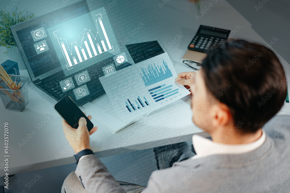 Poster individual analyzes financial data on laptop while reviewing printed reports and using smartphone in