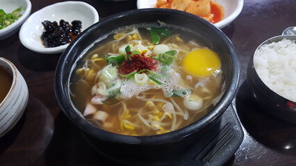 A traditional Korean bean sprout soup (kongnamul gukbap) served in a hot stone bowl with a raw egg, chili powder, and green onions. Accompanied by rice and assorted Korean side dishes.