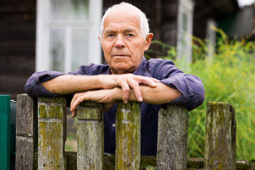 Portrait of pensive elderly man at fence of his country house in village