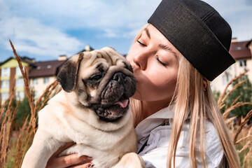Young happy beautiful woman holding, kissing and hugging her beloved dog during a walk with harness...