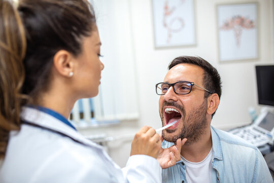 A female doctor examines a male patient&rsquo;s throat using a tongue depressor. The patient keeps his mouth open while the doctor inspects his tonsils and throat area.