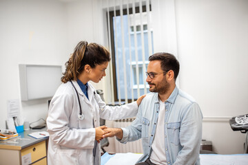 A female doctor holds a male patient's hand, offering reassurance and support during a clinical...