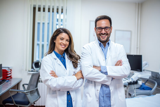 Portrait of two confident doctors standing in a modern medical office, smiling with arms crossed. Professional healthcare workers in white lab coats showcasing teamwork and trust in a clinical