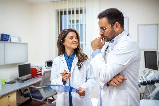 Two doctors in white coats have a thoughtful discussion in a bright medical office. The female doctor is holding a clipboard while the male doctor listens attentively.