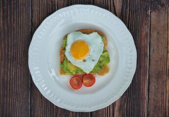Top view of Heart shaped fried egg on smashed avocado and tomato on a white plate on a wooden background