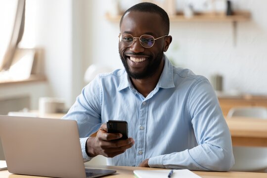 Happy african american man using smartphone and laptop at desk in home office light - Powered by Adobe