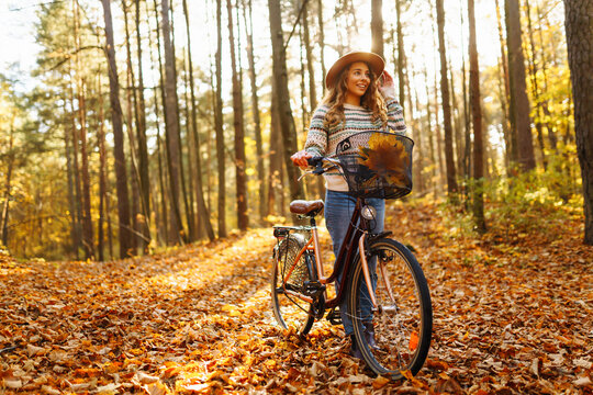 A cheerful woman with a bicycle rides through an autumn sunny forest surrounded by bright foliage. A young woman in a hat has fun and walks outdoors with a bicycle. The concept of fun, enjoyment.