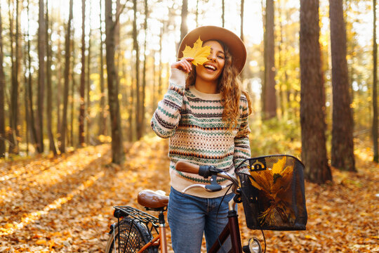 A cheerful woman with a bicycle rides through an autumn sunny forest surrounded by bright foliage. A young woman in a hat has fun and walks outdoors with a bicycle. The concept of fun, enjoyment.