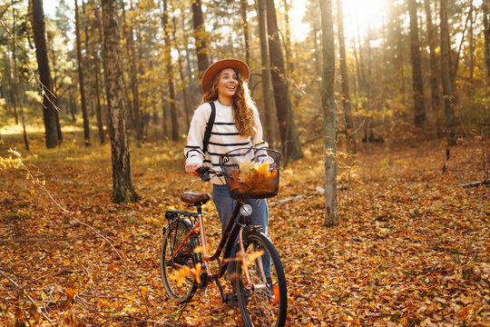 A cheerful woman with a bicycle rides through an autumn sunny forest surrounded by bright foliage. A young woman in a hat has fun and walks outdoors with a bicycle. The concept of fun, enjoyment.